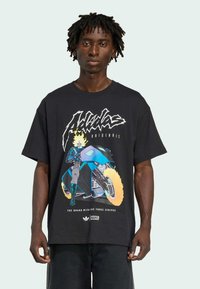 Ung mand iført en sort graphic T-shirt med et billede af en flammende kraniumrytter på en futuristisk blå motorcykel med Marvel- og Adidas-logoer.