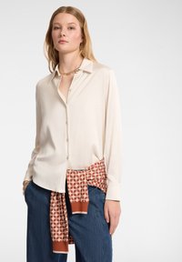 Camicia button-up color crema con colletto, abbinata a pantaloni blu e una sciarpa arancione con motivo legata in vita. Tessuto complessivamente liscio.