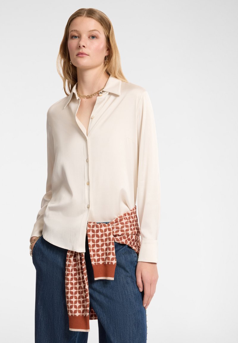 Camicia button-up color crema con colletto, abbinata a pantaloni blu e una sciarpa arancione con motivo legata in vita. Tessuto complessivamente liscio.