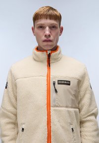 Cream-colored fleece jacket with orange zipper, two zip pockets, and logo patch. Features a high collar and textured fabric.
