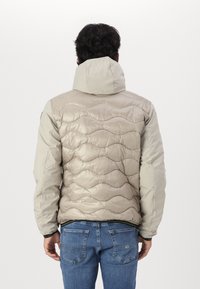 Giacca imbottita beige con cappuccio, caratterizzata da un motivo a onde e una finitura lucida sul retro, abbinata a jeans di denim blu.