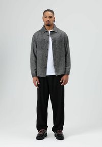 Grijze visgraatjas met twee borstzakken, gedragen over een wit overhemd. Zwarte losse broek en stevige zwarte schoenen met roze veters.