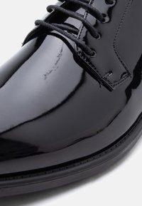 Κοντινό πλάνο γυαλιστερού μαύρου δερμάτινου παπουτσιού φόρεμα με μαύρα κορδόνια και λεπτομερή ραφή σε λευκό φόντο.