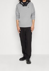 Pullover grigio con mezzo zip, rifiniture nere e logo. Abbinato a pantaloni neri e stivali neri, mostrando una vestibilità comoda e uno stile informale.