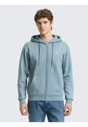 Junger Mann trägt einen hellblauen Kapuzenpullover mit Reißverschluss, Fronttaschen und Kordeln, kombiniert mit blauen Jeans, steht vor einem einfarbigen Hintergrund.