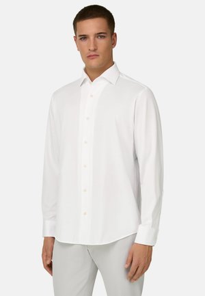 Man draagt een wit overhemd met lange mouwen en knoopmanchetten, gecombineerd met lichtgrijze broek, staand tegen een effen achtergrond.