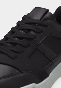 Gros plan sur une basket noire avec des lacets noirs, un panneau latéral texturé et une semelle blanche.