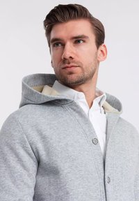 Cardigan grigio con cappuccio e chiusura a bottone, dotato di una morbida fodera interna e una finitura texture sull'esterno.