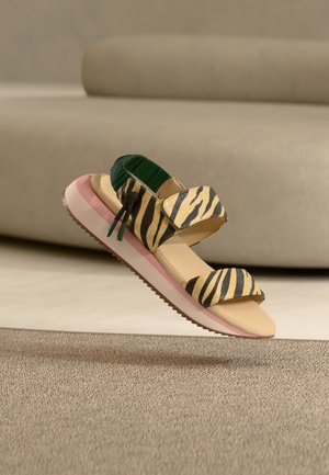 Sandale sportive avec des brides à rayures tigre, talon élastique vert, semelle beige avec une bande rose, présentée en suspension au-dessus d'un tapis beige.