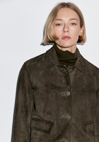 Olijfgroene suède jas met een kraag, knoopsluiting aan de voorkant, twee borstzakken en een zachte, matte textuur.