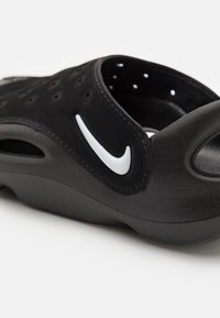 Musta kumisaapas, jossa on reikiä, kaareva muoto ja tyylikäs valkoinen Nike-swoosh-logo sivulla. Sujuva pinta ja pyöreä kärki.