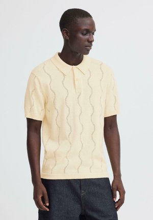 Man draagt een lichtgele korte mouw gebreide poloshirt met een zigzagpatroon, gecombineerd met donkerblauwe spijkerbroek.