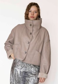 Afgekorte taupekleurige leren jas met hoge kraag, voorzien van knoopsluitingen en zachte, met bont gevoerde manchetten. Draagt erbij zilverkleurige broeken met slangenpatroon.