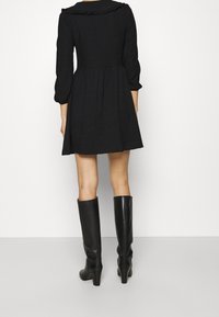 Svart klänning med långa ärmar och volangkrage; utsvängd kjol. Matchad med knähöga svarta stövlar med klack. Slätt tyg, enkel design.