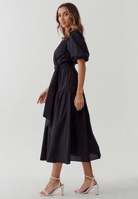 Zwarte katoenen midi-jurk met puffmouwen, een ceintuur in de taille en een gelaagde rok. Gecombineerd met witte sandaaltjes met hakken.