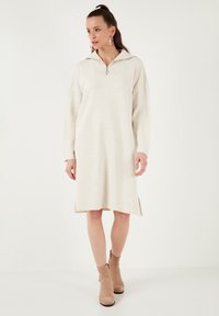 Beiges Strickkleid mit halbem Reißverschlusskragen, langen Ärmeln und seitlichen Schlitzen. Weicher, strukturierter Stoff, knielang, kombiniert mit beigen Knöchelstiefeln.