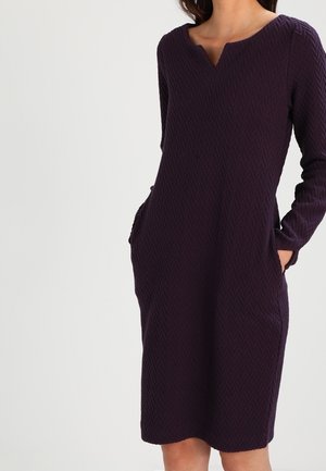 Kobieta w ciemnofioletowej sukience o długości do kolan, z długim rękawem, fakturowanej, z dekoltem w wycięcie i rękami w kieszeniach bocznych.