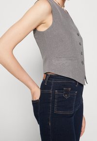 En person klädd i en grå knappad väst och mörkblå jeans med orange sömmar, med handen vilande i det främre fickan.