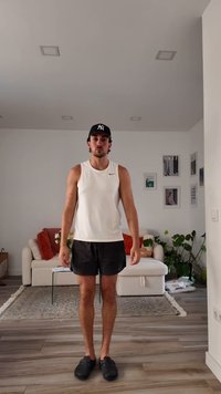 Man wearing black cap, white sleeveless shirt, black shorts, and black shoes standing in a living room with sofa and plants.