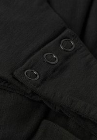 Tejido negro con una superficie ligeramente texturizada que presenta tres botones redondos negros en el puño, ofreciendo un diseño limpio y minimalista.