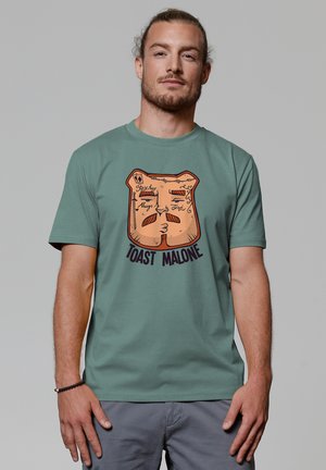 T-shirt à manches courtes, vert menthe, avec un graphisme de pain grillé en dessin animé, le texte "TOAST MALONE," et des accents en orange et noir.