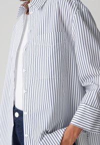 Camicia bianca con bottoni, caratterizzata da strisce verticali blu, una tasca sul petto e una vestibilità rilassata, con colletto e maniche con risvolto.