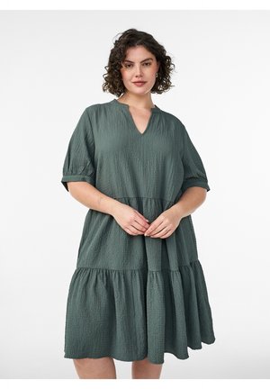 Frau mit lockigem Haar, die ein strukturiertes, locker sitzendes, knielanges grünes Kleid mit kurzen Ärmeln vor weißem Hintergrund trägt.