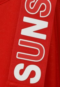 Roter Stoff mit einem weißen Druckdesign, das "SUNS" buchstabiert. Der Text ist fett und vertikal entlang eines Riemens positioniert.