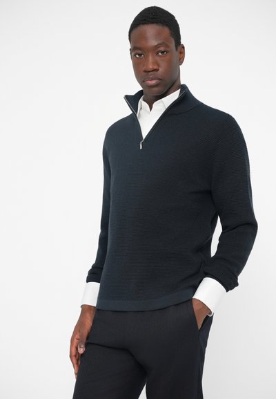 Pulover în nuanță închisă de bleumarin, cu guler tip quarter-zip și tricot texturat. Are manșete albe care ies de sub mâneci, asortate cu pantaloni închiși la culoare.