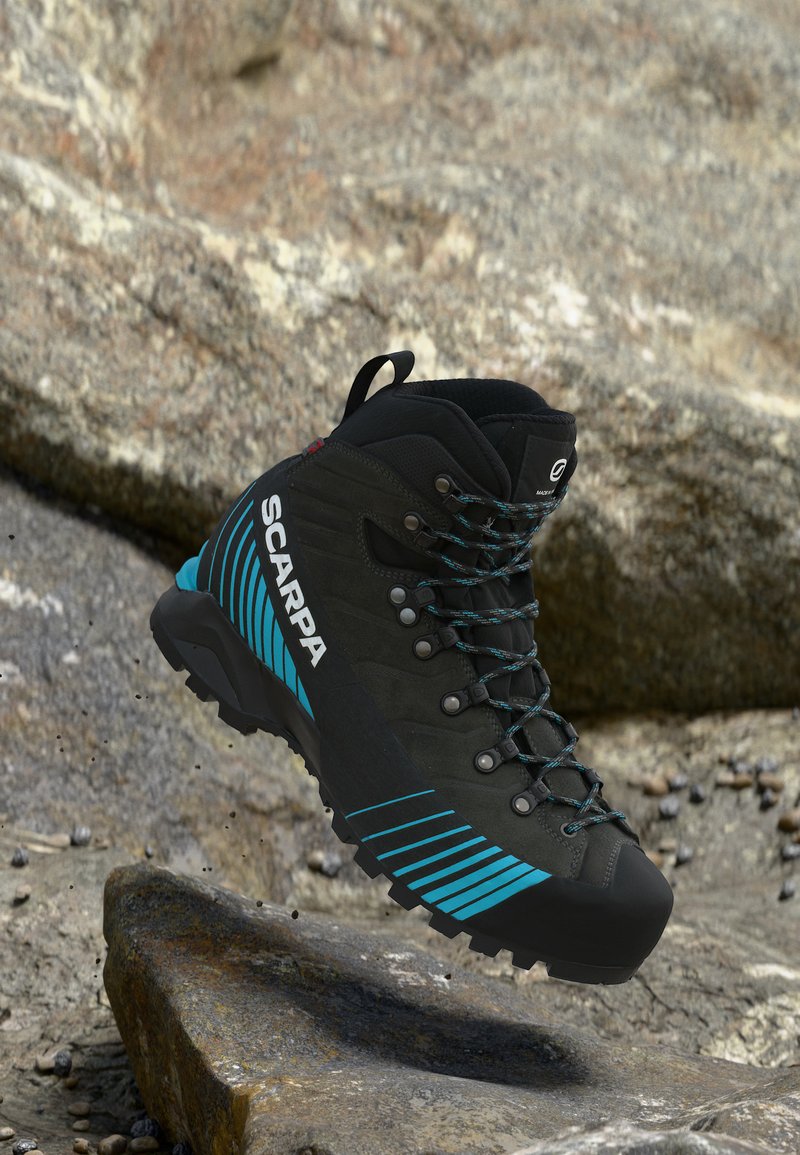 Bota de trekking negra con rayas azules y el logo SCARPA flotando sobre un terreno rocoso.