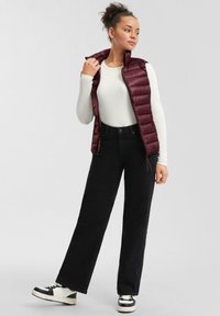 Junge Frau mit lockigem Haar, die ein weißes Langarmoberteil, eine kastanienbraune Steppweste, schwarze weite Hose und schwarz-weiße Turnschuhe trägt.