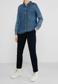 Denimová košile na knoflíky ve světle modré barvě s náprsní kapsou, spárovaná s námořnickými kalhotami a bílými teniskami. Košile má dlouhé rukávy a límec.