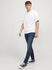Un jeune homme marche, portant un t-shirt blanc uni, un jean skinny bleu foncé et des baskets noires à faible hauteur sur un fond blanc.
