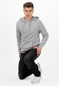 Felpa grigia con tasca frontale e coulisse, abbinata a jeans neri strappati e sneakers bianche, con dettagli rossi sulla suola.
