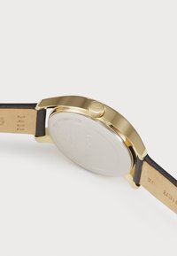 Reloj dorado con caja redonda, correa de cuero negra y fondo de metal plateado liso. Presenta una corona en el lado para ajustar la hora.