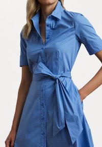 Ljust blå skjortklänning med korta ärmar, krage, knäppning framtill och detalj med knyt i midjan. Slät textur och utsvängd kjol med fickor.