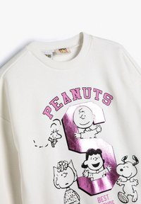 Biała bluza z różowym napisem "PEANUTS" oraz błyszczącą różową cyfrą 3, przedstawiająca postacie z kreskówki: Charlie Brown, Lucy, Sally, Snoopy i Woodstock.