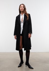 Femme portant un long manteau noir sur une chemise blanche à boutons et un short noir, vêtue de chaussettes noires jusqu'aux genoux et de talons noirs, se tenant sur un sol gris.