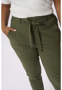 Personne portant un pantalon taille haute vert olive avec un lien à l'avant et la main dans la poche, associé à un haut blanc à manches courtes et un bracelet en or.