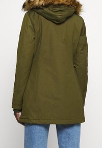 Person iført en olivengrøn parkajakke med hætte i pels, synlige sømme og lyseblå jeans, vist fra ryggen.