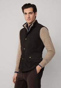 Gilet da uomo trapuntato marrone scuro con accessori dorati, dotato di due tasche frontali, colletto alto e un aspetto leggero abbinato a un maglione beige.
