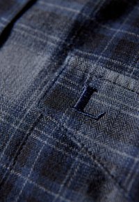 Blauw herringbone-patroon stof met een zak die een donkerblauwe geborduurde "L" heeft. Gestructureerd oppervlak, close-up opname die de details van de stof benadrukt.