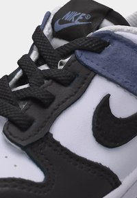Εγγύς λήψη αθλητικού παπουτσιού Nike με μαύρα κορδόνια, μαύρο λογότυπο swoosh, λευκές και μπλε δερμάτινες επιφάνειες και ορατό λογότυπο Nike στη γλώσσα.
