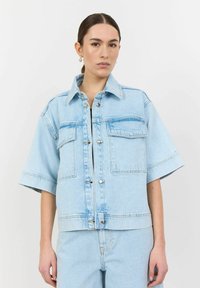 Svetlo modra denim jakna s kratkimi rokavi, gumbnim zapenjanjem in dvema žepoma na prsih. Ima klasičen ovratnik in sproščen kroj.