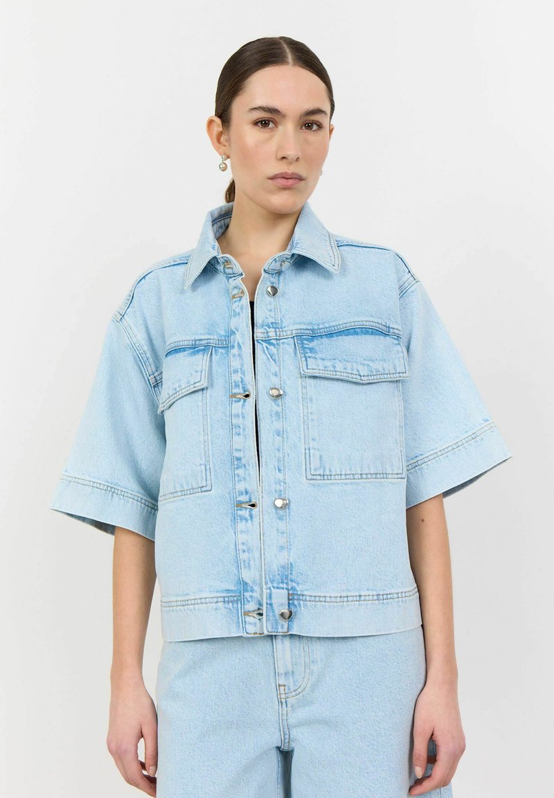 Svetlo modra denim jakna s kratkimi rokavi, gumbnim zapenjanjem in dvema žepoma na prsih. Ima klasičen ovratnik in sproščen kroj.