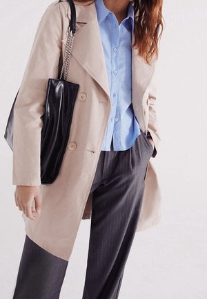 Osoba ubrana w beżowy płaszcz przeciwdeszczowy, jasnobłękitną koszulę zapinaną na guziki oraz szare spodnie w cienkie prążki, trzymająca czarną skórzaną torbę na ramię z łańcuszkowym paskiem.