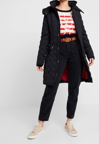 Černý prošívaný dlouhý kabát s vysokým límcem a patentkami, kombinovaný s červeně pruhovaným svetr, černými džíny a béžovými teniskami. Červená podšívka.