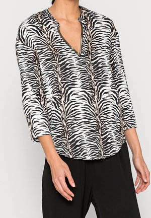 Femme portant un chemisier à col en V à rayures zèbre noir et blanc avec des manches trois-quarts et un pantalon noir.