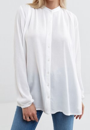 Woman wearing a long-sleeve, buttoned white blouse with a round collar and blue jeans, standing against a plain background.