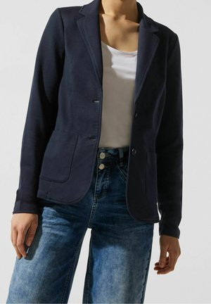 Kobieta ubrana w granatową marynarkę na białej bluzce, zestawioną z niebieskimi dżinsami z wysokim stanem, stojąca z rękoma swobodnie opuszczonymi wzdłuż ciała.